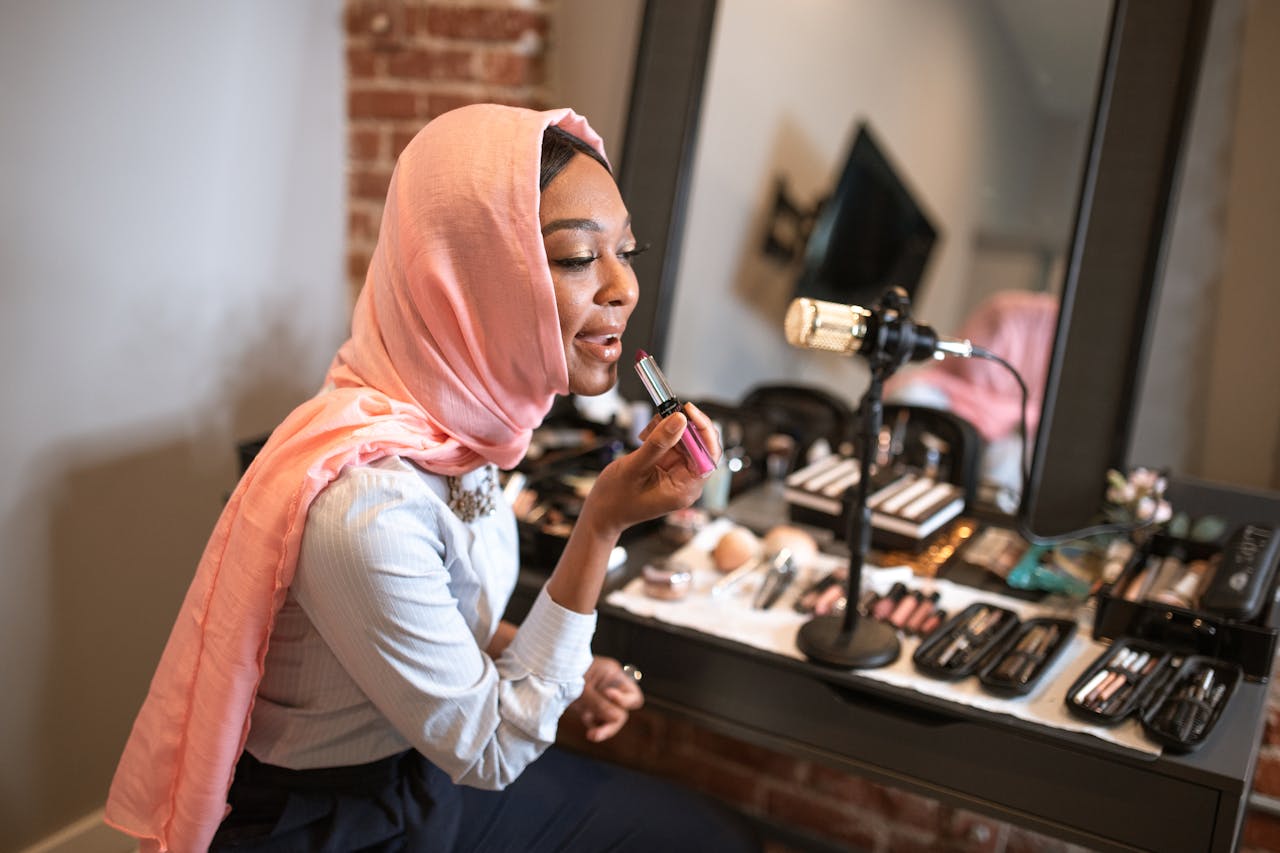 Muslim woman in pink hijab applying makeup while recording a beauty vlog indoors.